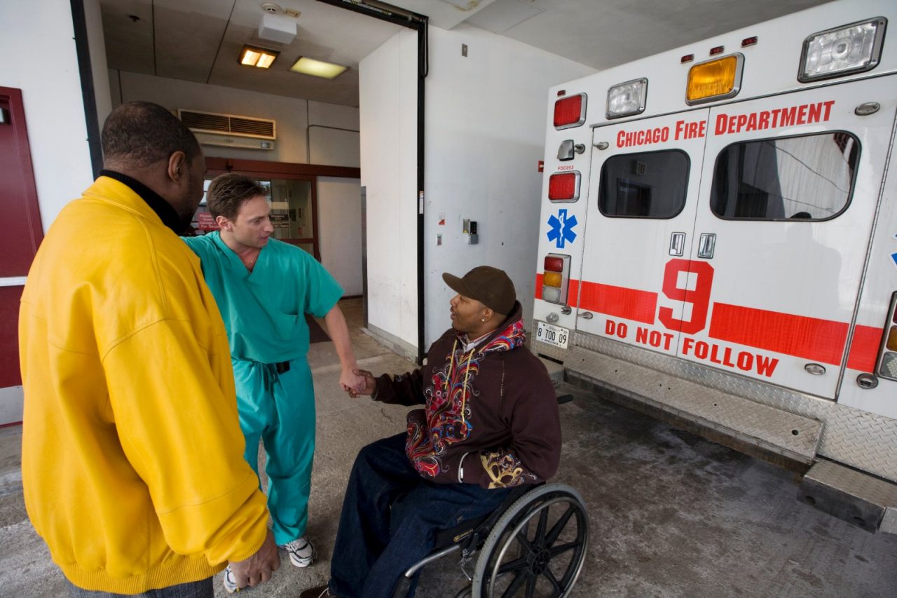 A doctor and another man talk with a man in a wheelchair, next to an ambulance.