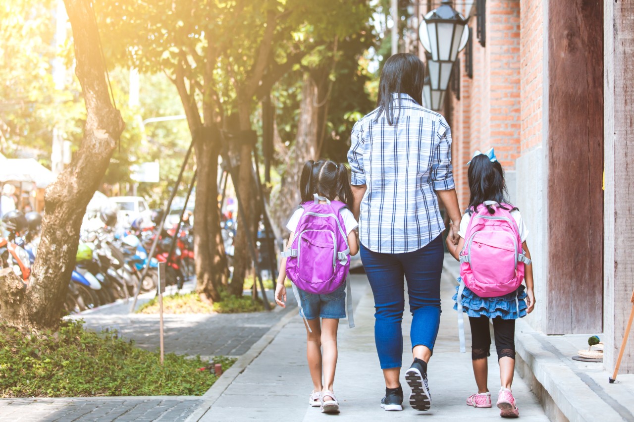 A mother and two young daughters with backpacks hold hands while walking on a sidewalk.