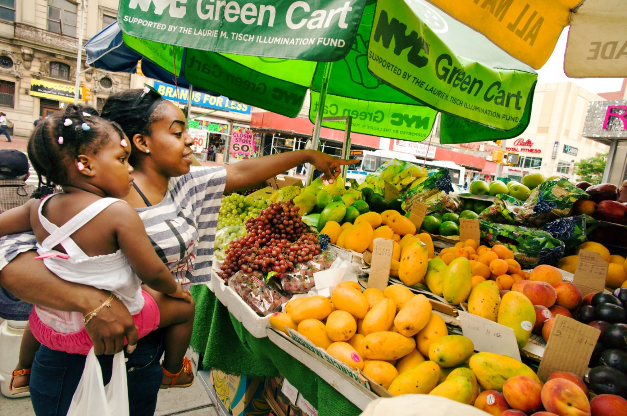 A woman and child pick fresh fruits and vegetables from a New York City Green Cart in Jamaica, Queens, NYC.