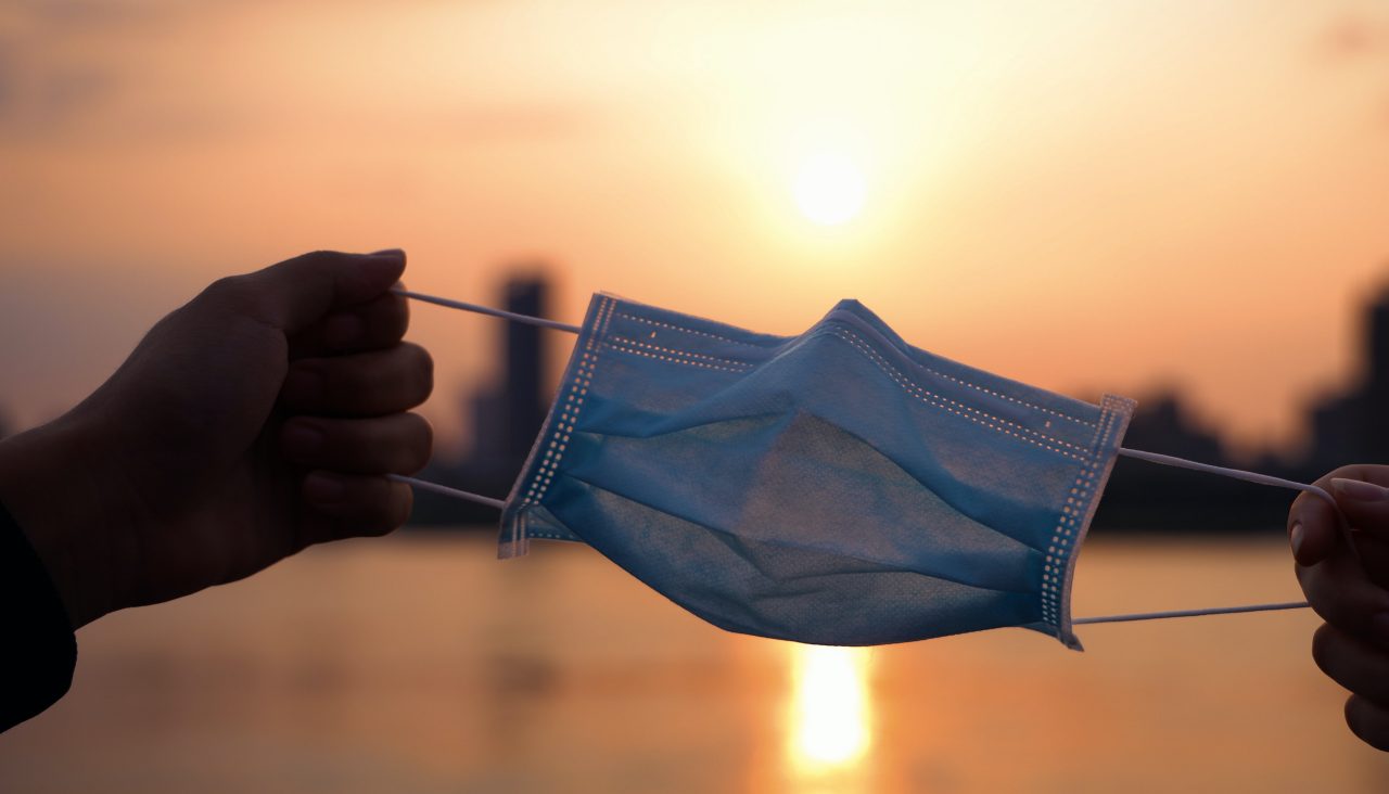 Two hands holding opposite ends of a mask in front of a sunset.