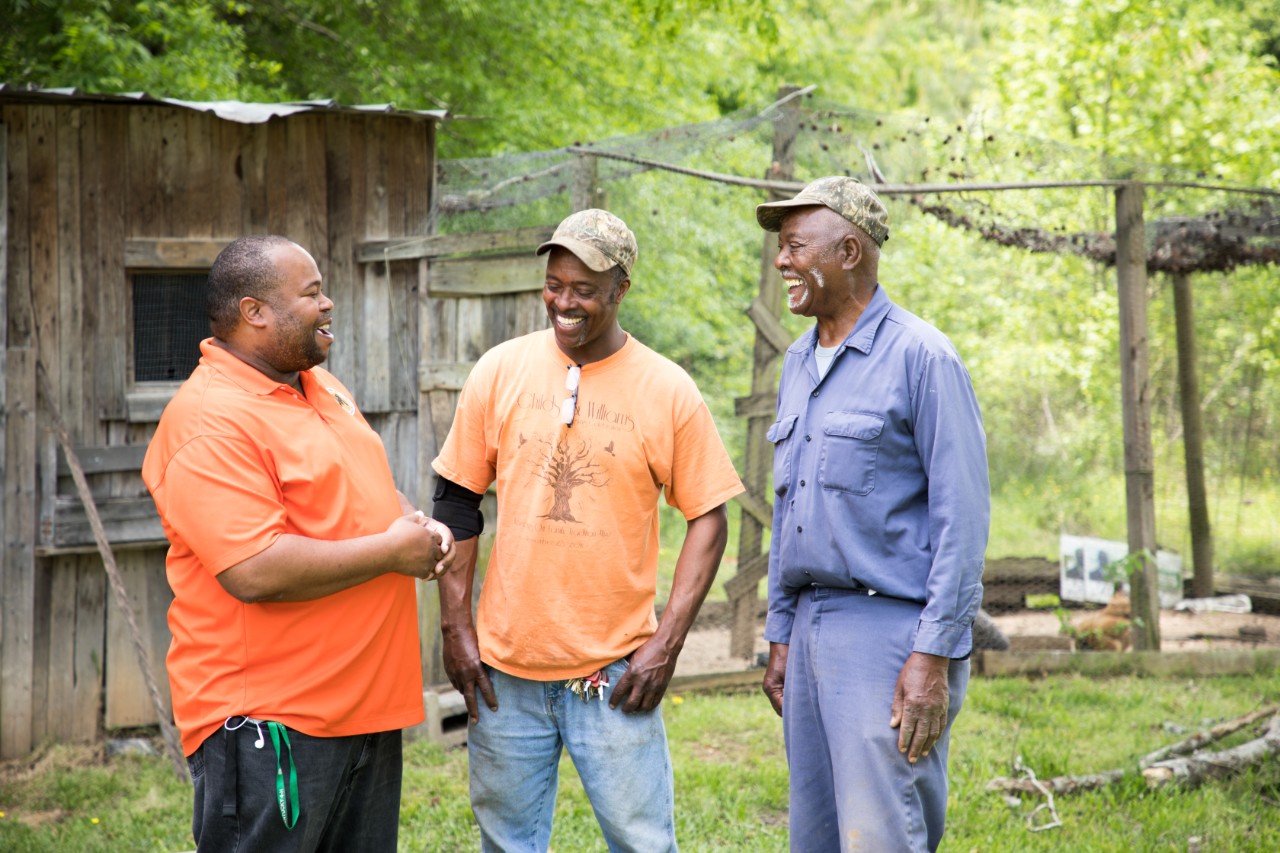 Three men smile while carrying on a conversation outside.