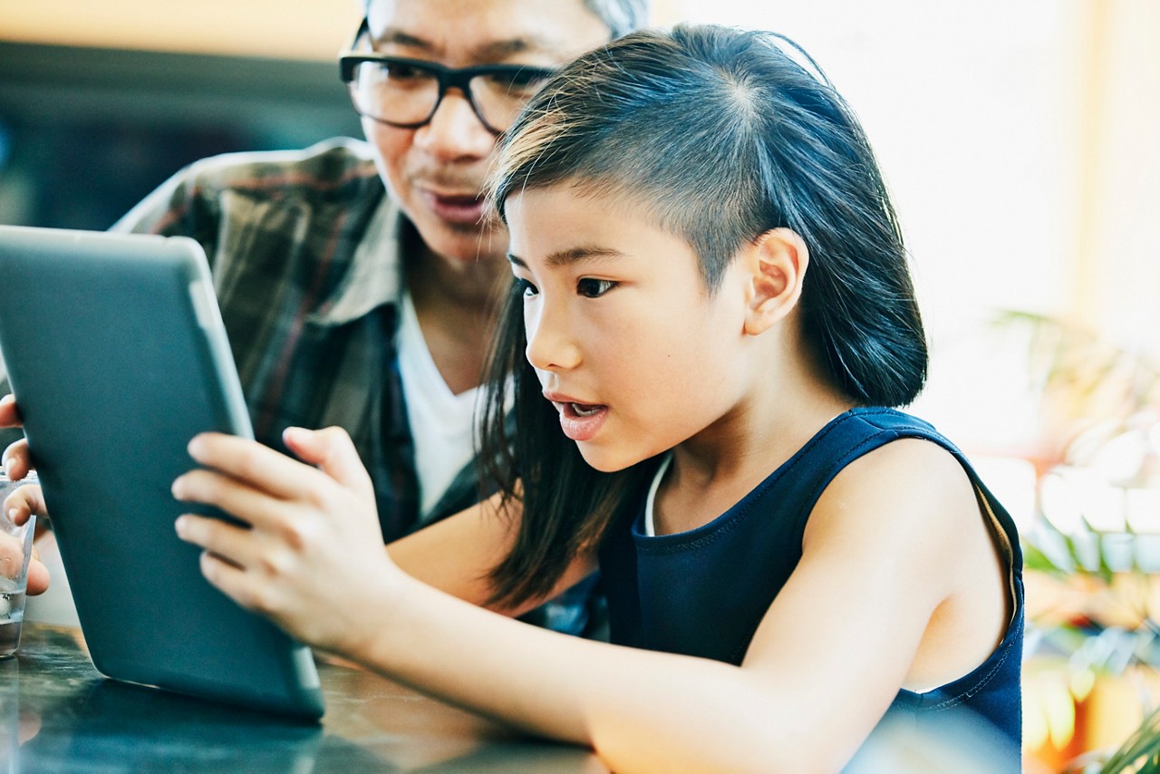 1053592394 Young girl looking at video on digital tablet with father at kitchen table