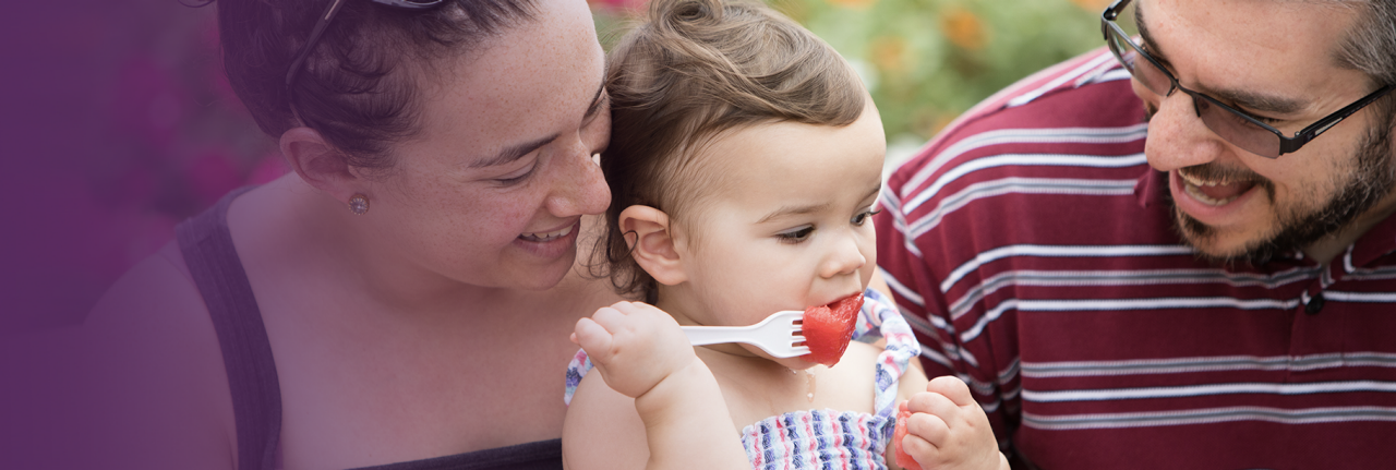 Improving Child and Family Wellbeing A mother and father smile at their toddler while she eats watermelon.