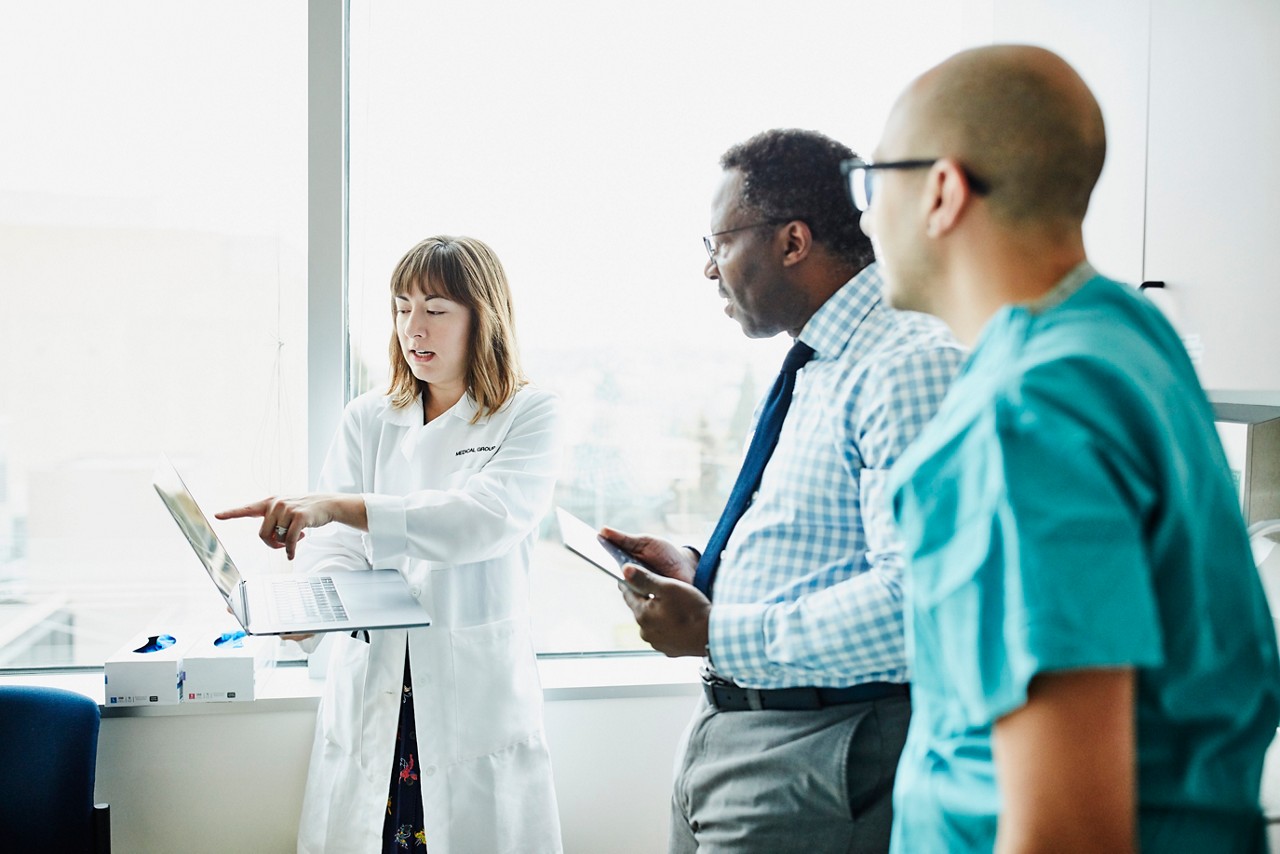 Female doctor holding laptop discussing patient information with medical team in exam room Female doctor holding laptop discussing patient information with medical team in exam room