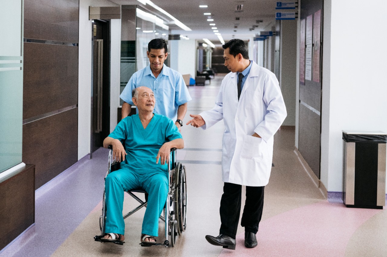 Male porter pushing man in his 70s along corridor, male doctor gesturing and talking