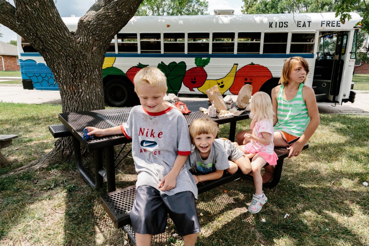 Iola, KS - August 1, 2017 - Local youth (L to R) Conner McCoullough, Ryan Cress, Lilly Cunningham, and Katlin Cress eat lunch at a picnic table in front of the MARV ("Meals And Reading Vehicle") bus in a residential neighborhood in Iola.