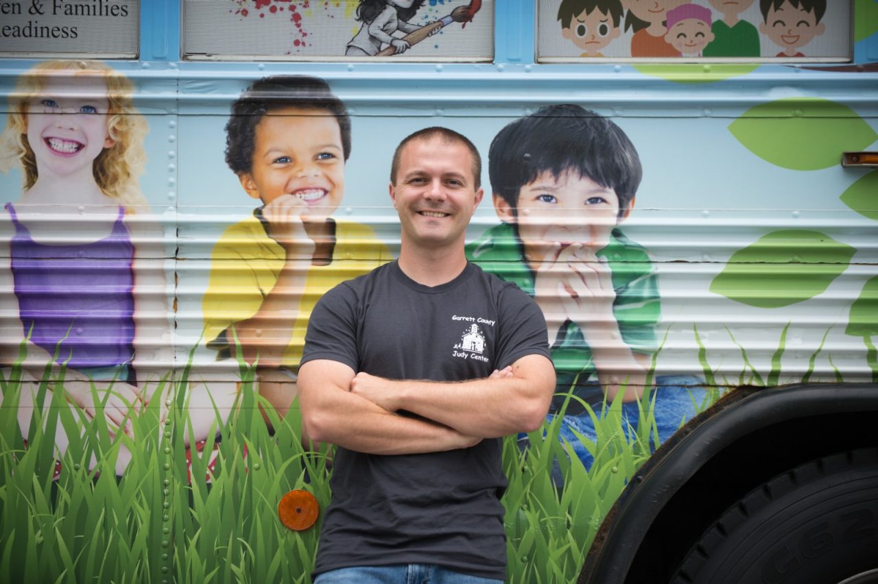 Steve Knepp stands next to the Learning Beyond the Classroom Bus in front of Crellin Elementary School. The bus holds a library and learning games for children, and offers healthy snacks.