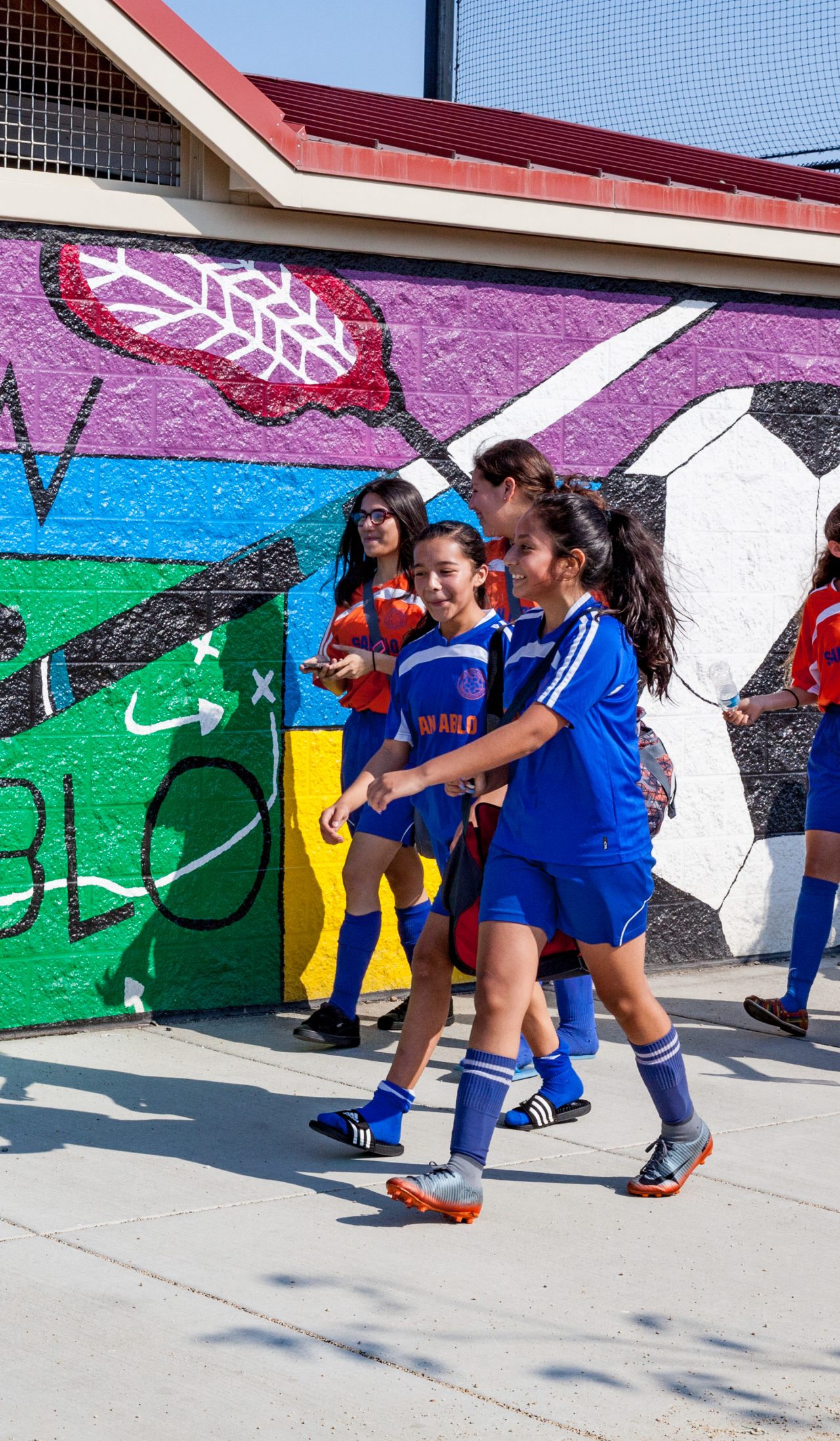 A girls' soccer team walks by an outdoor wall mural. 