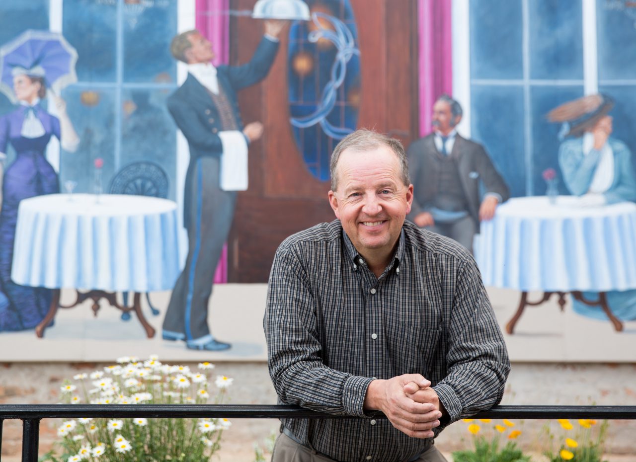 A man leaning on a railing, with a mural of people dining in the background.