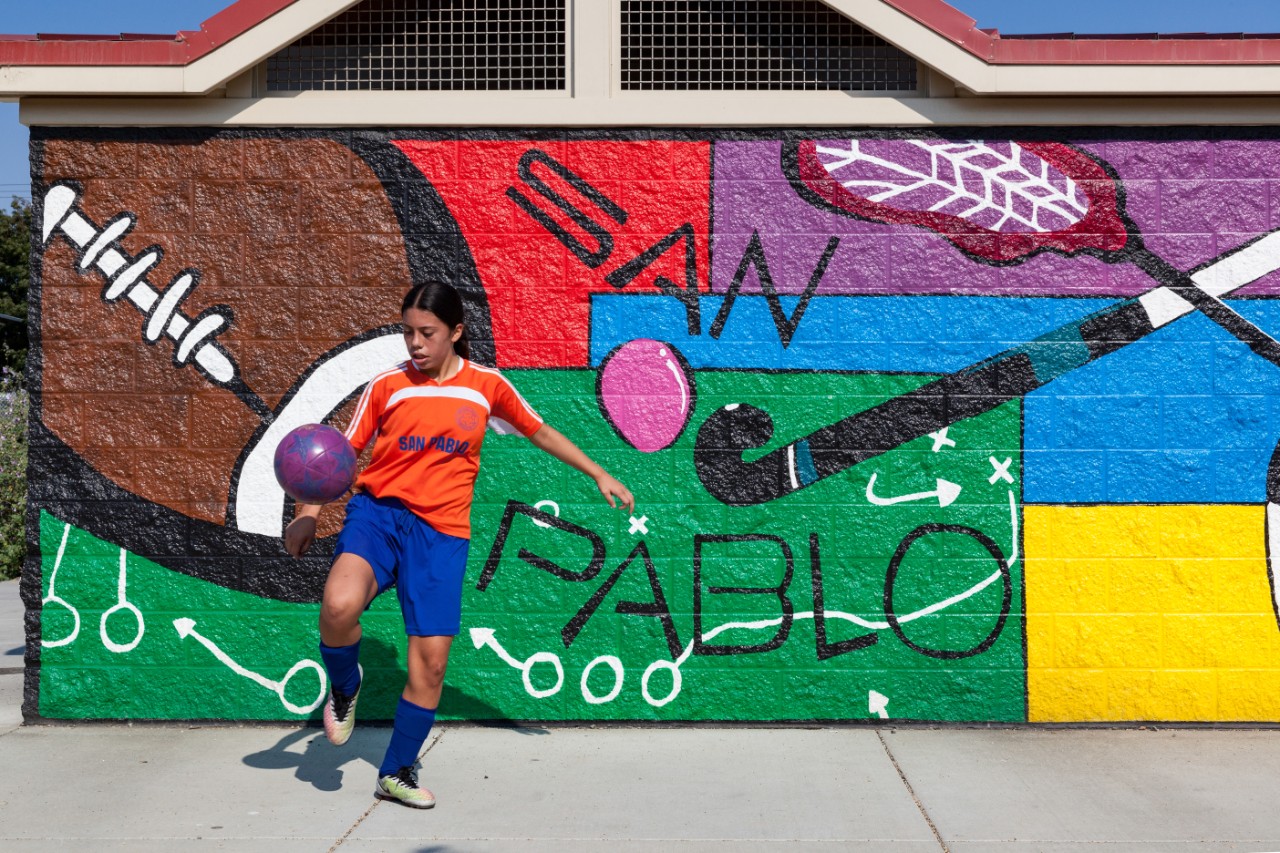 A youth soccer player juggles a ball in front of a mural highlighting sports equipment.