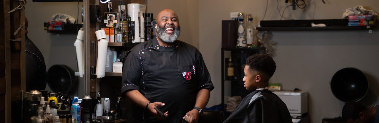 A barber smiles as he prepares to cut a young man’s hair.