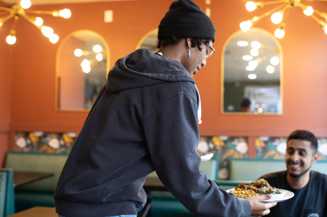 A Black woman serves a meal to a customer seated at a table. 
