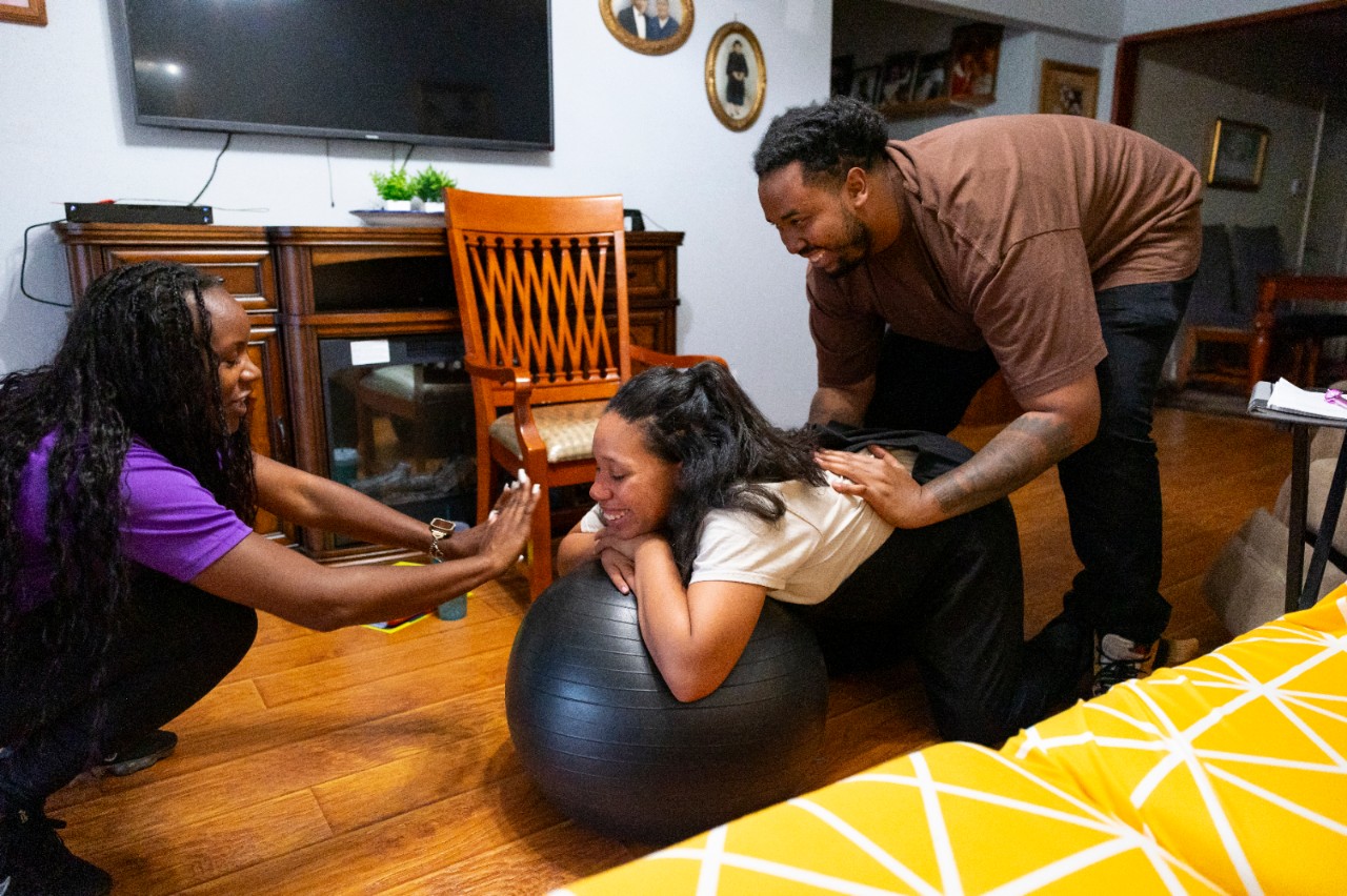 A doula instructs a pregnant woman and a man standing behind her while the woman leans over a birthing ball.