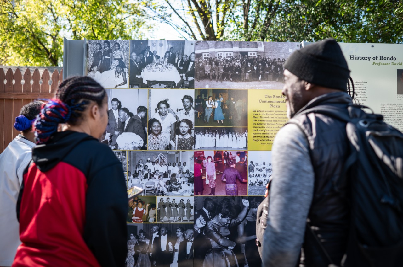 Two people standing looking at a board with black and white photos, the board reads, “History of Rondo.” 