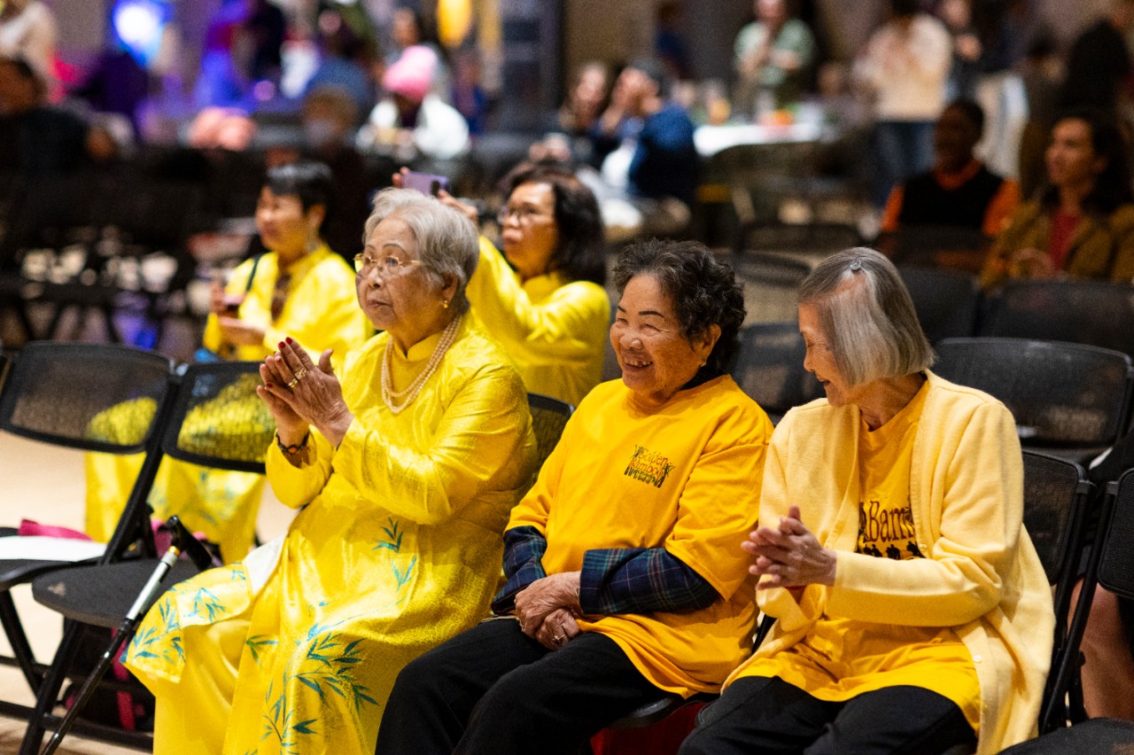 Three people in yellow clothing sit and clap for a performance.