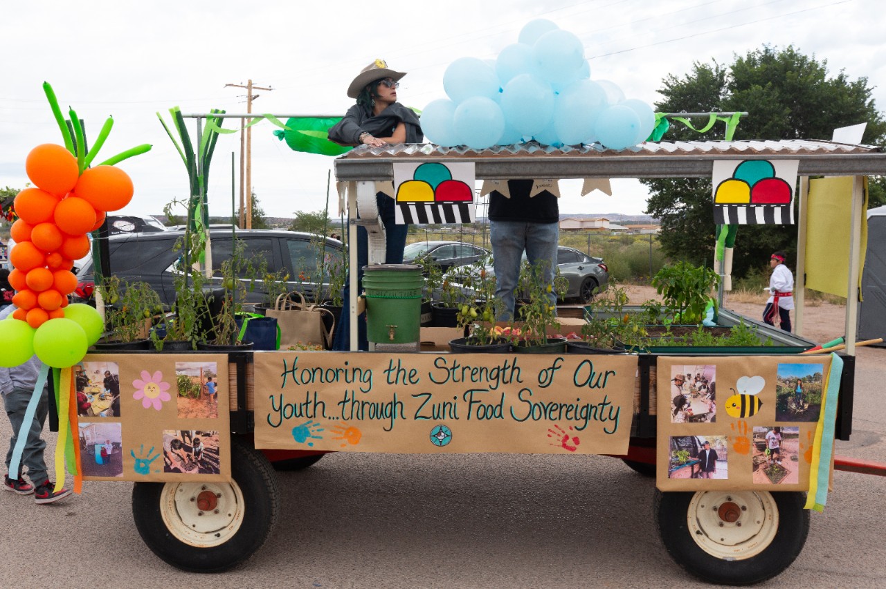 A float parade cart decorated with ballons in the shape of corn and peas. Different plants sit on the cart and there is one person standing on top. Decorative signs read: “honoring the strength of our youth through Zuni food sovereignty.” 