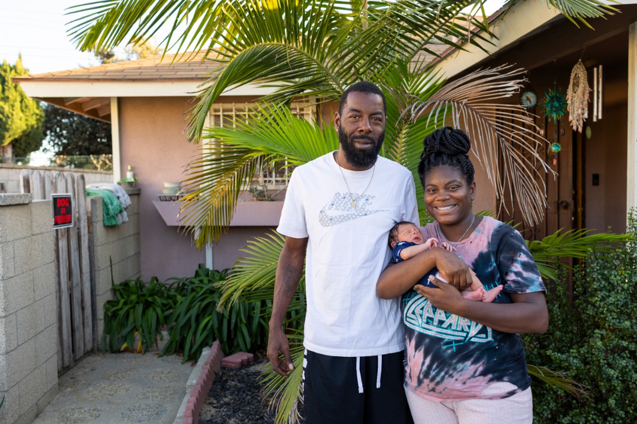 Two parents stand in front of their home in Los Angeles holding their newborn baby. 