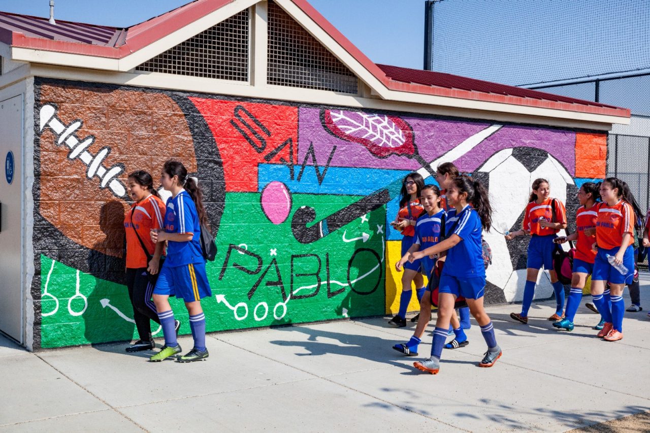 A girls' soccer team walks by an outdoor wall mural. 
