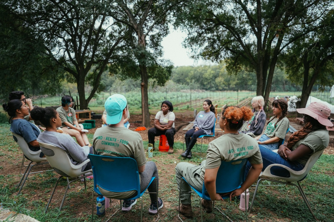 Members of a youth group sit outside, in a circle, with a vegetable garden in the background.