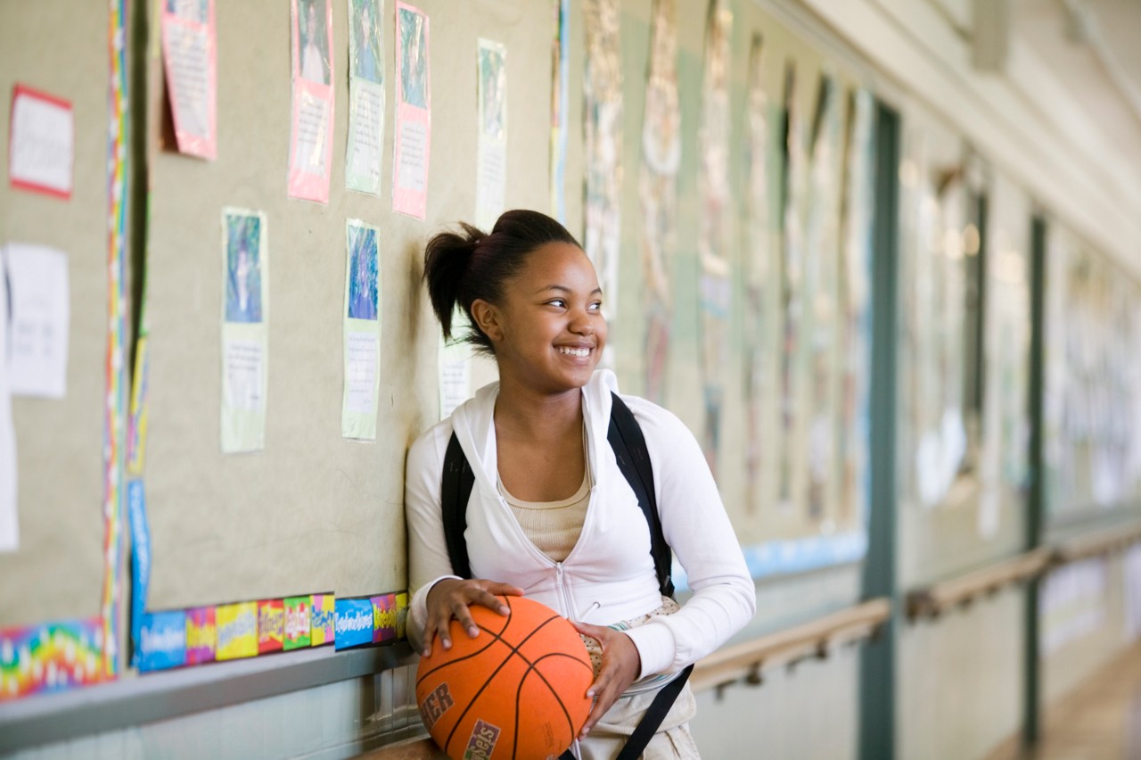 A smiling student holds a basketball while standing in a school hallway. A girl with a basketball at her school. Sports 4 Kids (Playworks)