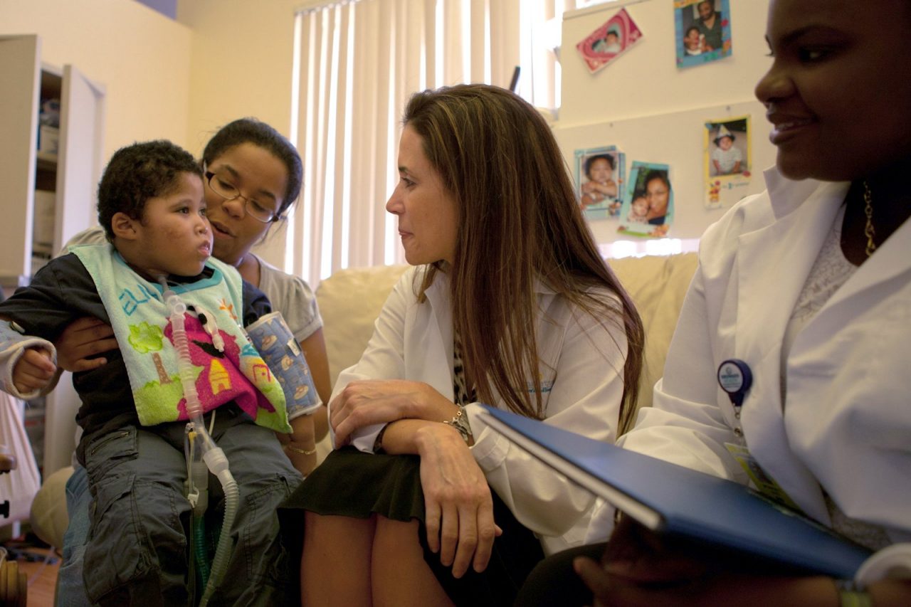 Medical professionals talk with a disabled child who sits on his mother's lap.