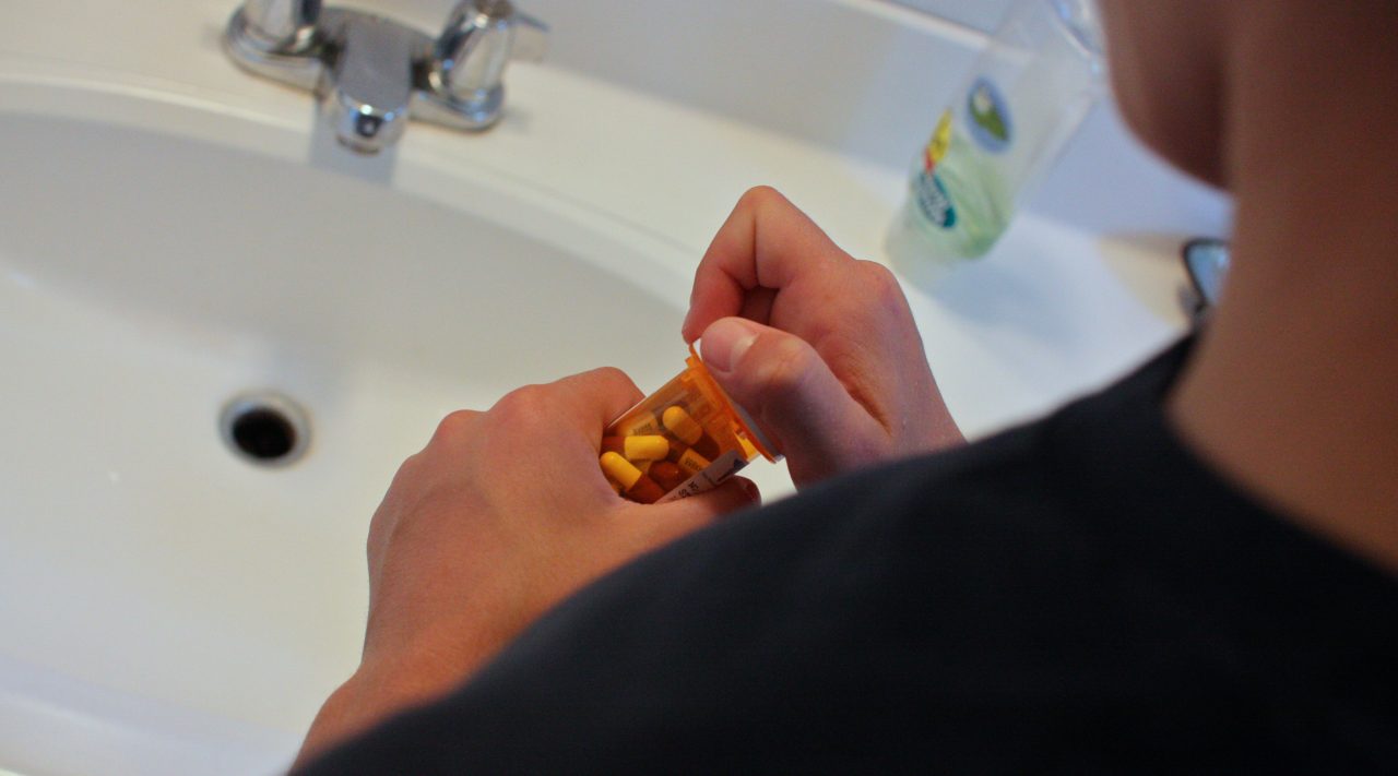 A boy standing by a sink, opening a bottle of pills.