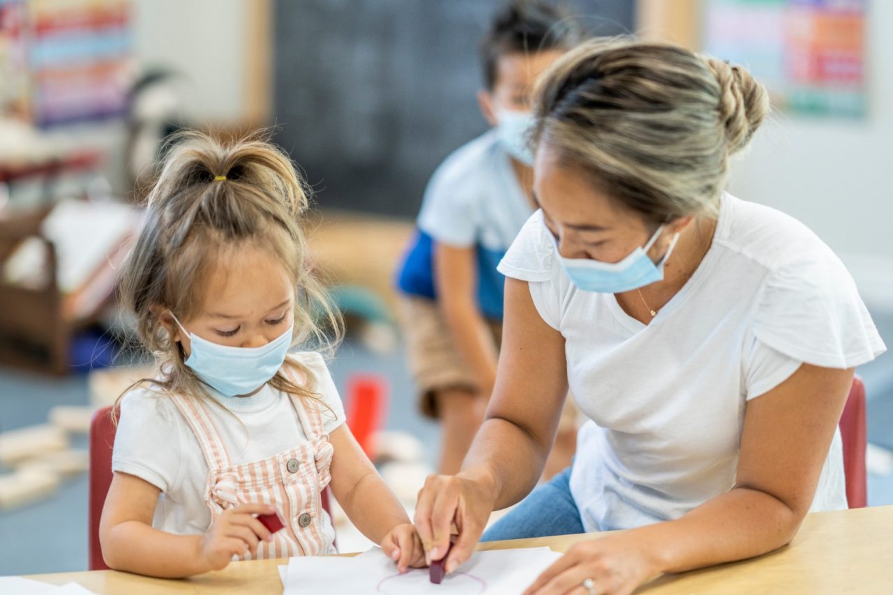 Cute young girl colouring in a daycare facility while wearing a protective face mask to avoid the transfer of germs during COVID-19.
