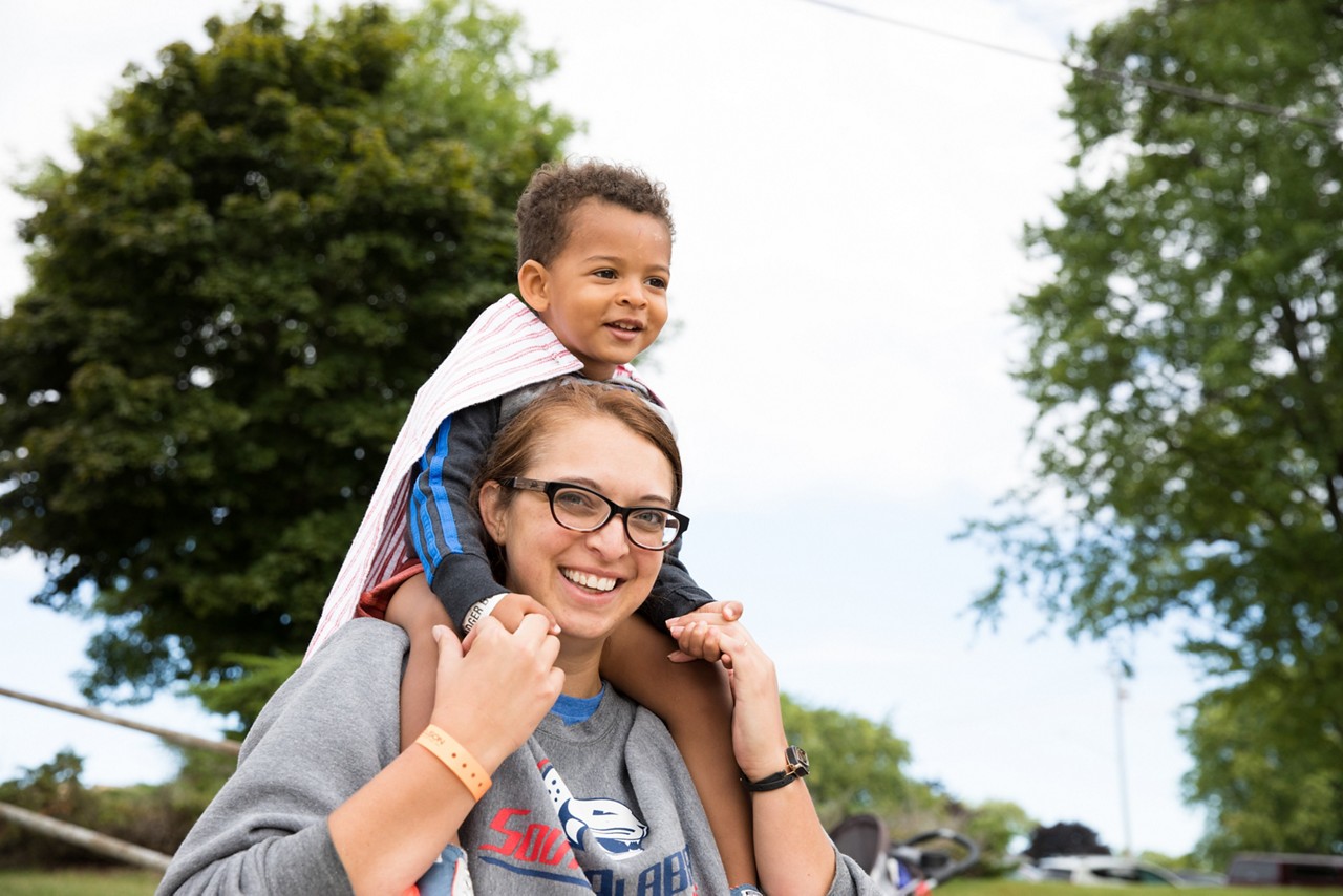 A smiling mother carries her child on her shoulders outside.