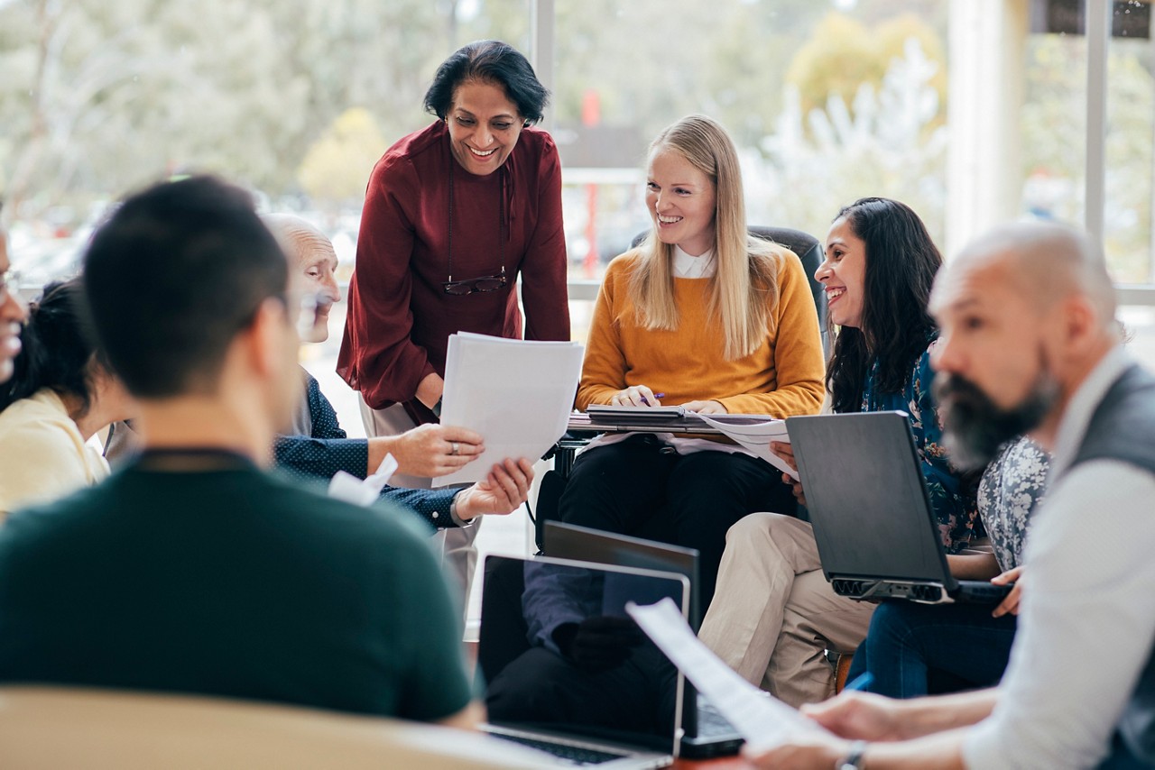 A diverse team looks happy in conversation. They are sitting around with laptops in front of them. Woman of Indian ethnicity leads the team A diverse team looks happy in conversation. They are sitting around with laptops in front of them. Woman of Indian ethnicity leads the team