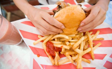 Hands of woman holding hamburger, with fries and ketchup. A strawberry milkshake is on the side.