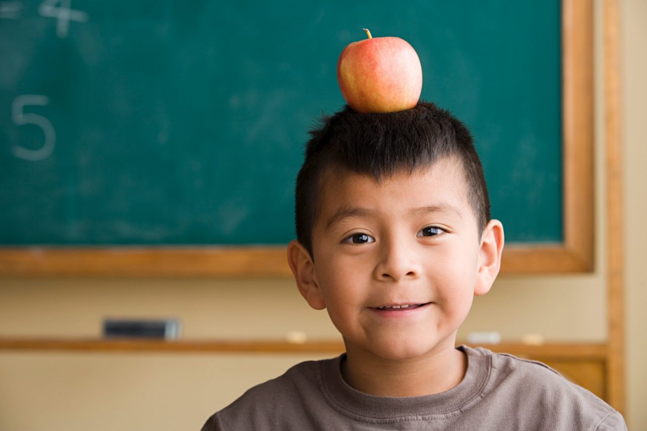A boy standing in front of a chalkboard, balancing an apple on his head.