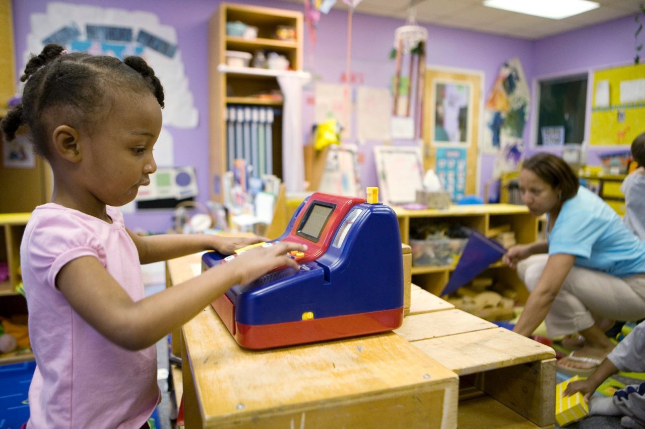 Young girl playing with make believe cash register. A teacher is in the background. Nariya Farrington playing on a toy cash register in her childcare center classroom. Frank Porter Graham Child Development Center, Chapel Hill, North Carolina.