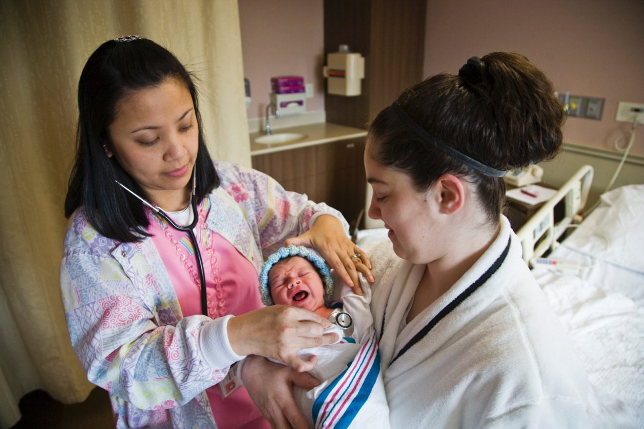 A nurse checks a crying newborn baby's temperature and heartbeat, Mom holds the baby and watches.  Hackensack University Medical Center in Hackensack, NJ.


