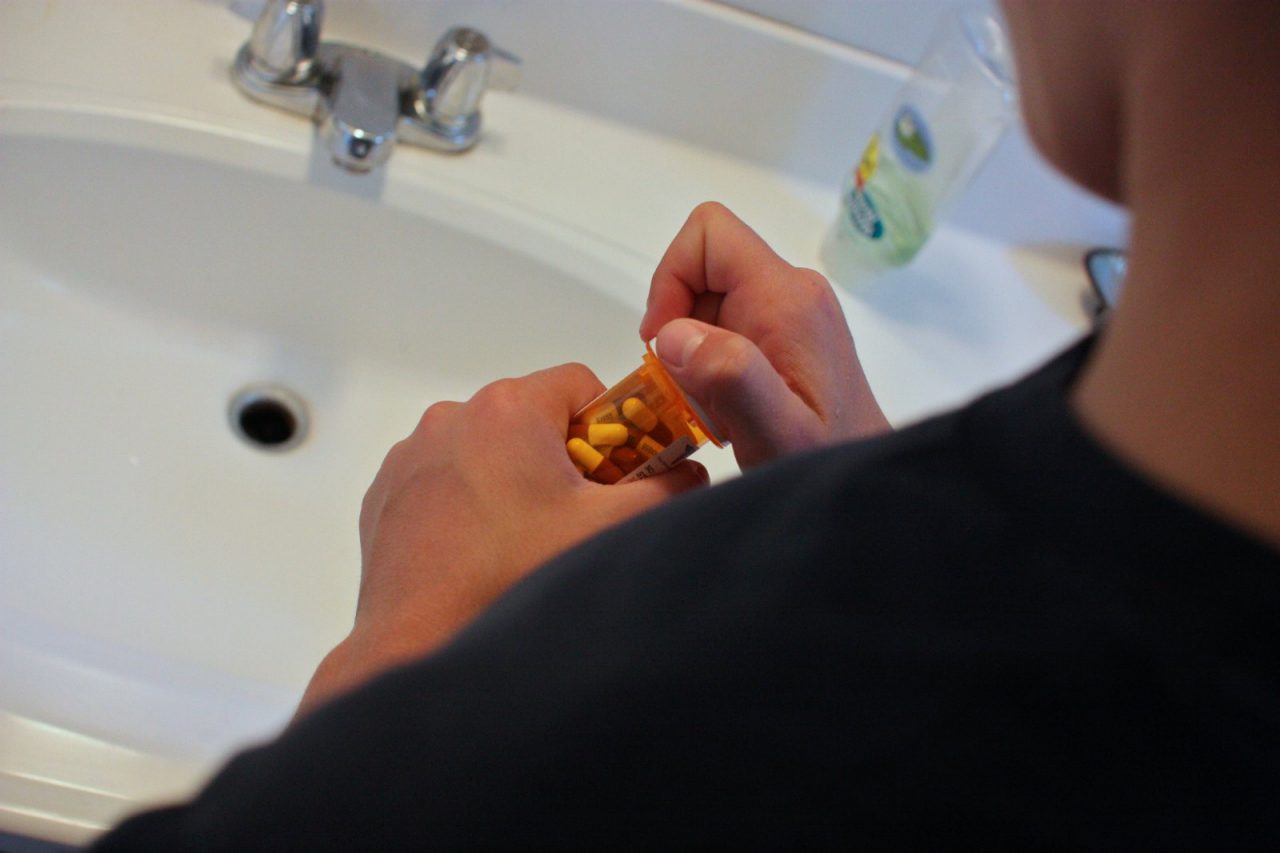 A boy standing by a sink, opening a bottle of pills.