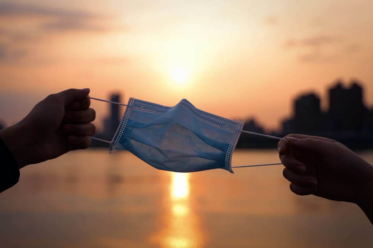 Two hands holding opposite ends of a mask in front of a sunset.