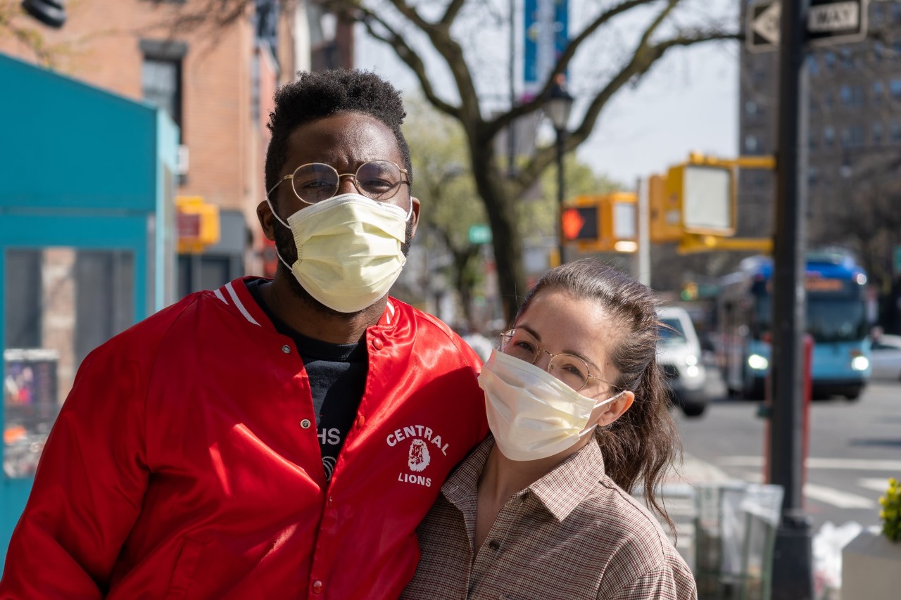A man and woman stand on a sidewalk wearing masks.