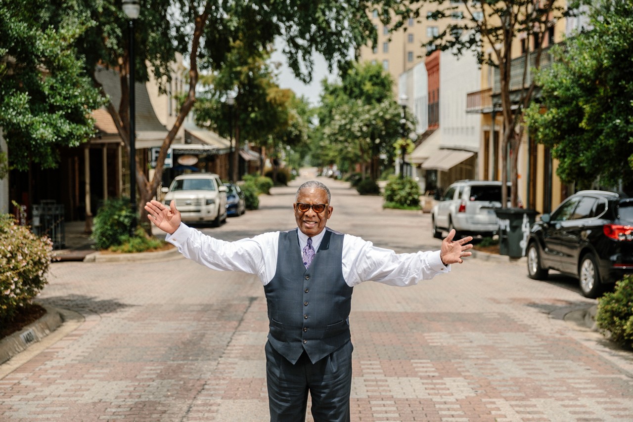 A man, wearing a suit, stands open armed in the middle of a downtown street. 