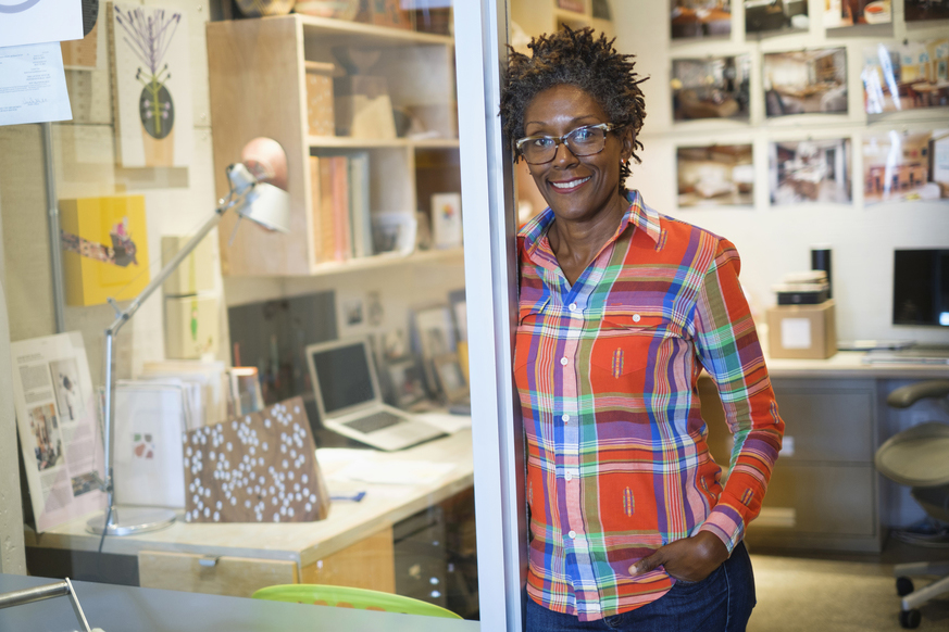 One black woman in casual, colorful shirt, standing in doorway of office.