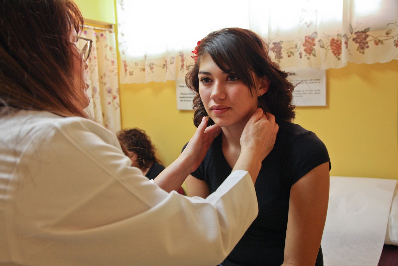 A doctor examines a teenage patient, her mother sits in the background, Hidalgo Medical Services-Bayard Community Health Center, Bayard, New Mexico. Project ECHO.