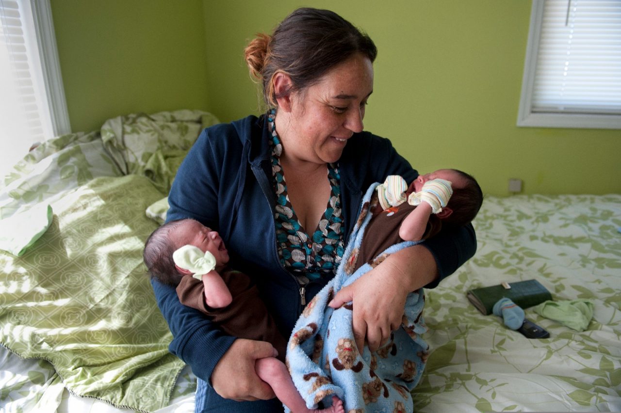 A smiling mother holding her infant twins, while sitting in her bedroom.