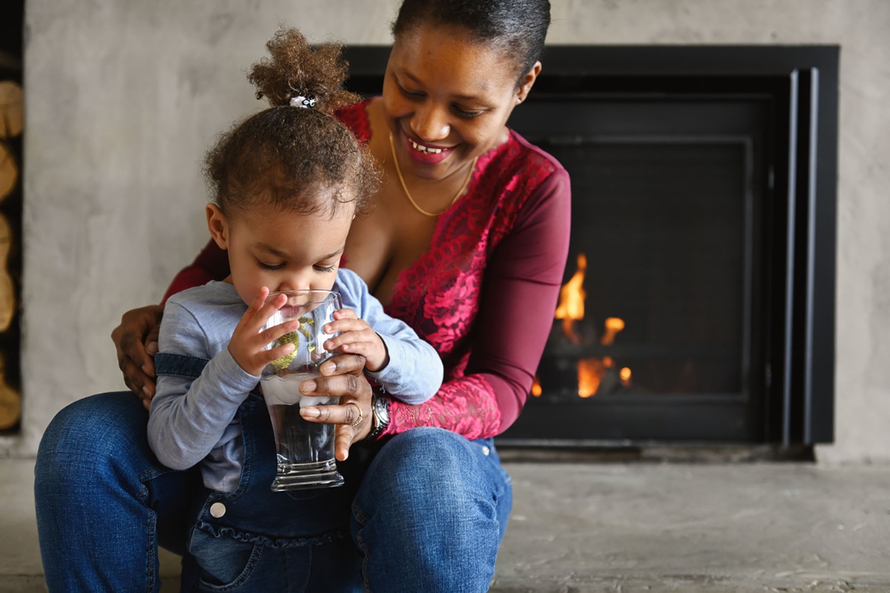 A mother sitting with young child drinking water in front of fireplace.