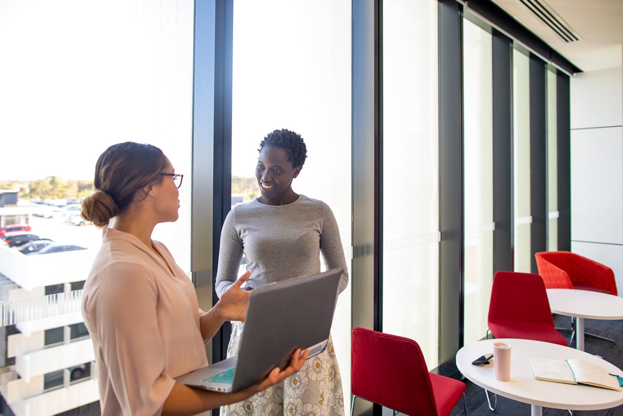 Two women standing by large windows together while using a laptop open in front of one of them.