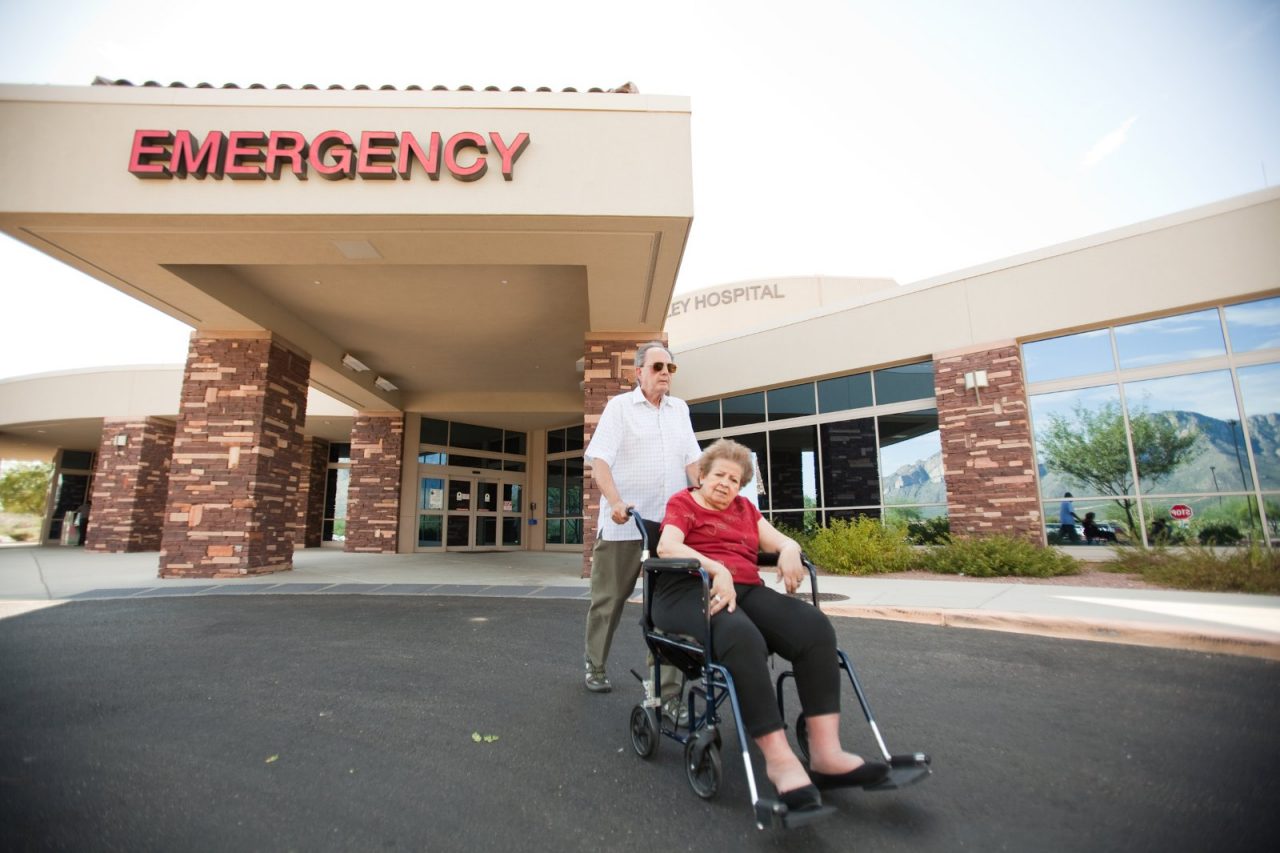An elderly man wheels his wheelchair bound wife away from a hospital emergency wing.