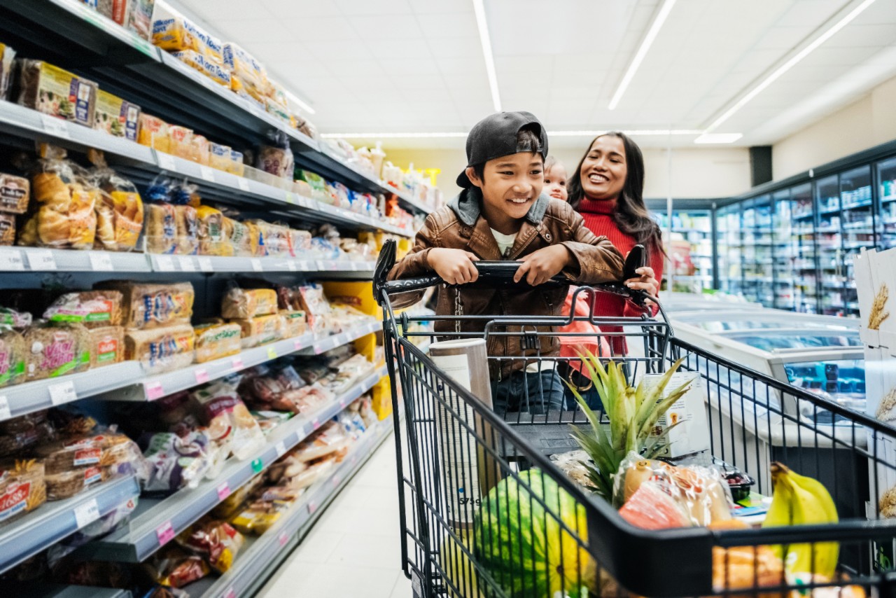 A young boy having fun on a shopping cart while out buying groceries with his mother.